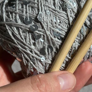 May include: A ball of light gray yarn interwoven with small, reflective silver sequins. Two smooth, light-colored bamboo knitting needles are positioned diagonally across the yarn, resting against the textured surface of the ball.