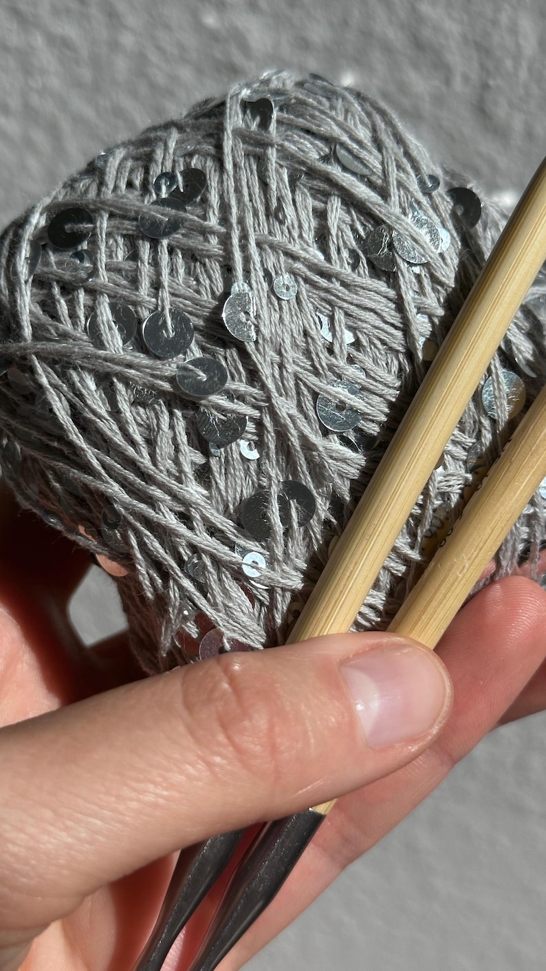 May include: A ball of light gray yarn interwoven with small, reflective silver sequins. Two smooth, light-colored bamboo knitting needles are positioned diagonally across the yarn, resting against the textured surface of the ball.