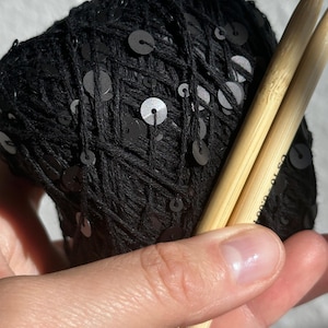 May include: A ball of black yarn featuring integrated black sequins, paired with two light-colored bamboo knitting needles. The needles have metallic silver tips and clearly display the text US 10 - 6.00 mm printed on the side.