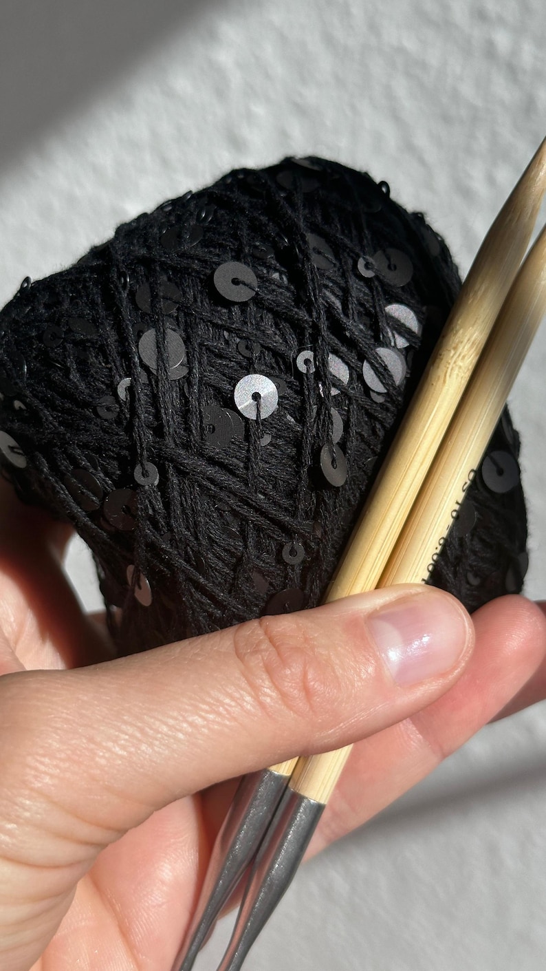 May include: A ball of black yarn featuring integrated black sequins, paired with two light-colored bamboo knitting needles. The needles have metallic silver tips and clearly display the text US 10 - 6.00 mm printed on the side.