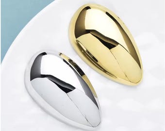 May include: Two smooth, teardrop-shaped metal objects with a highly reflective, polished finish. One object is gold-toned, and the other is silver-toned. Both pieces feature a rounded, convex surface that catches light, creating distinct highlights.