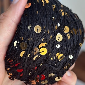 May include: A ball of black yarn featuring small, round, metallic gold sequins attached throughout the thread. The yarn is wound into a compact, rounded shape, showcasing the contrast between the dark fiber and the reflective gold accents.