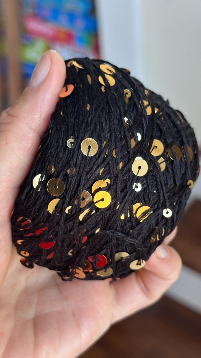 May include: A ball of black yarn featuring small, round, metallic gold sequins attached throughout the thread. The yarn is wound into a compact, rounded shape, showcasing the contrast between the dark fiber and the reflective gold accents.