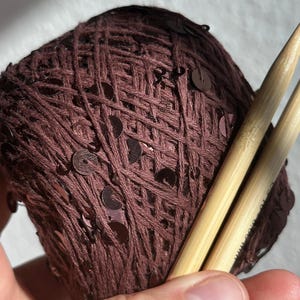 May include: A ball of dark brown yarn interwoven with small, matching brown sequins. Two light-colored bamboo knitting needles are inserted into the yarn ball, with the text 8.00 mm clearly visible on the surface of one of the needles.