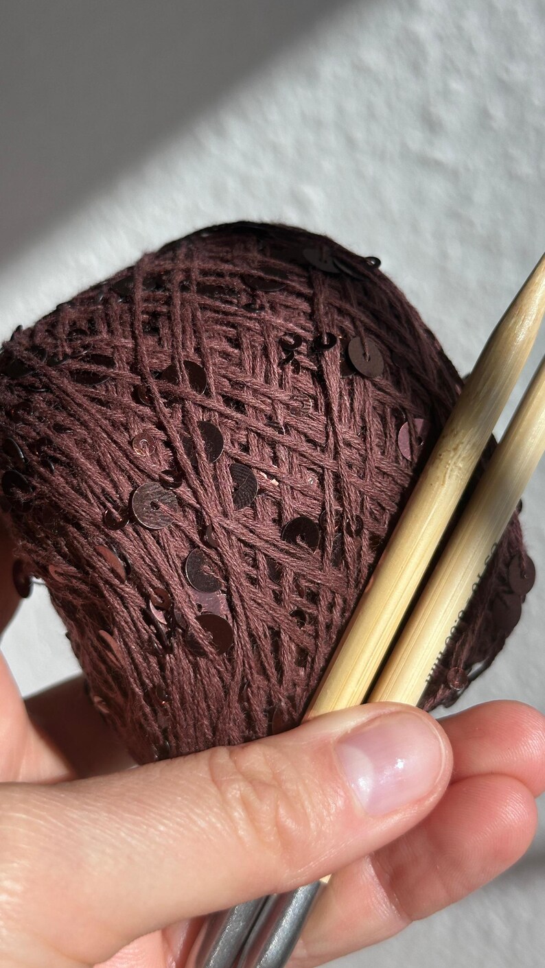 May include: A ball of dark brown yarn interwoven with small, matching brown sequins. Two light-colored bamboo knitting needles are inserted into the yarn ball, with the text 8.00 mm clearly visible on the surface of one of the needles.