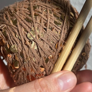 May include: A ball of brown yarn interwoven with reflective gold sequins, paired with two smooth, light-colored bamboo knitting needles. The yarn features a consistent texture throughout the ball, and the metallic sequins are distributed evenly across the surface, catching the light to create a bright, shimmering effect against the matte brown fibers.