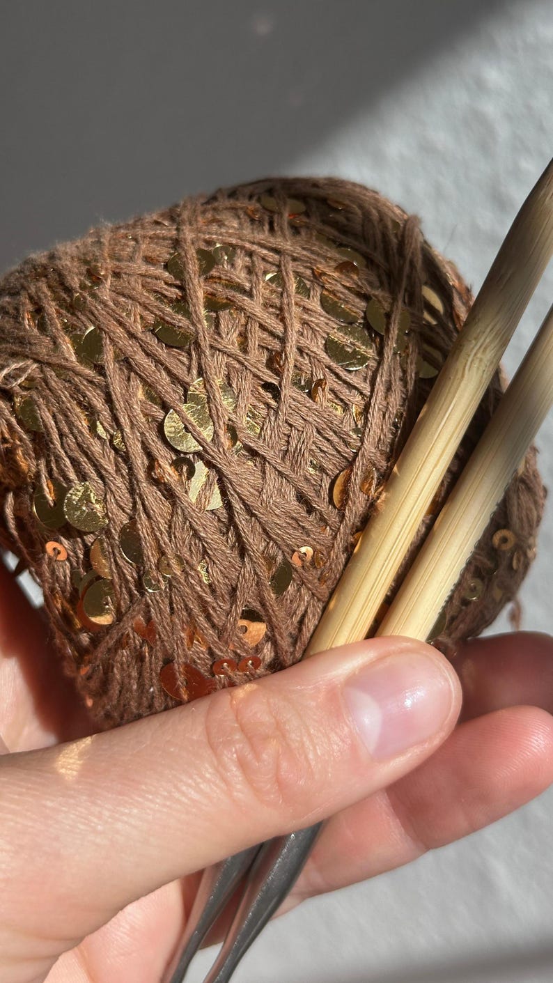 May include: A ball of brown yarn interwoven with reflective gold sequins, paired with two smooth, light-colored bamboo knitting needles. The yarn features a consistent texture throughout the ball, and the metallic sequins are distributed evenly across the surface, catching the light to create a bright, shimmering effect against the matte brown fibers.