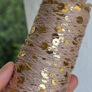 May include: A cylindrical spool of light brown yarn wrapped with small, reflective gold sequins. The metallic gold discs are distributed across the surface of the wound thread, creating a shimmering texture throughout the entire length of the spool.