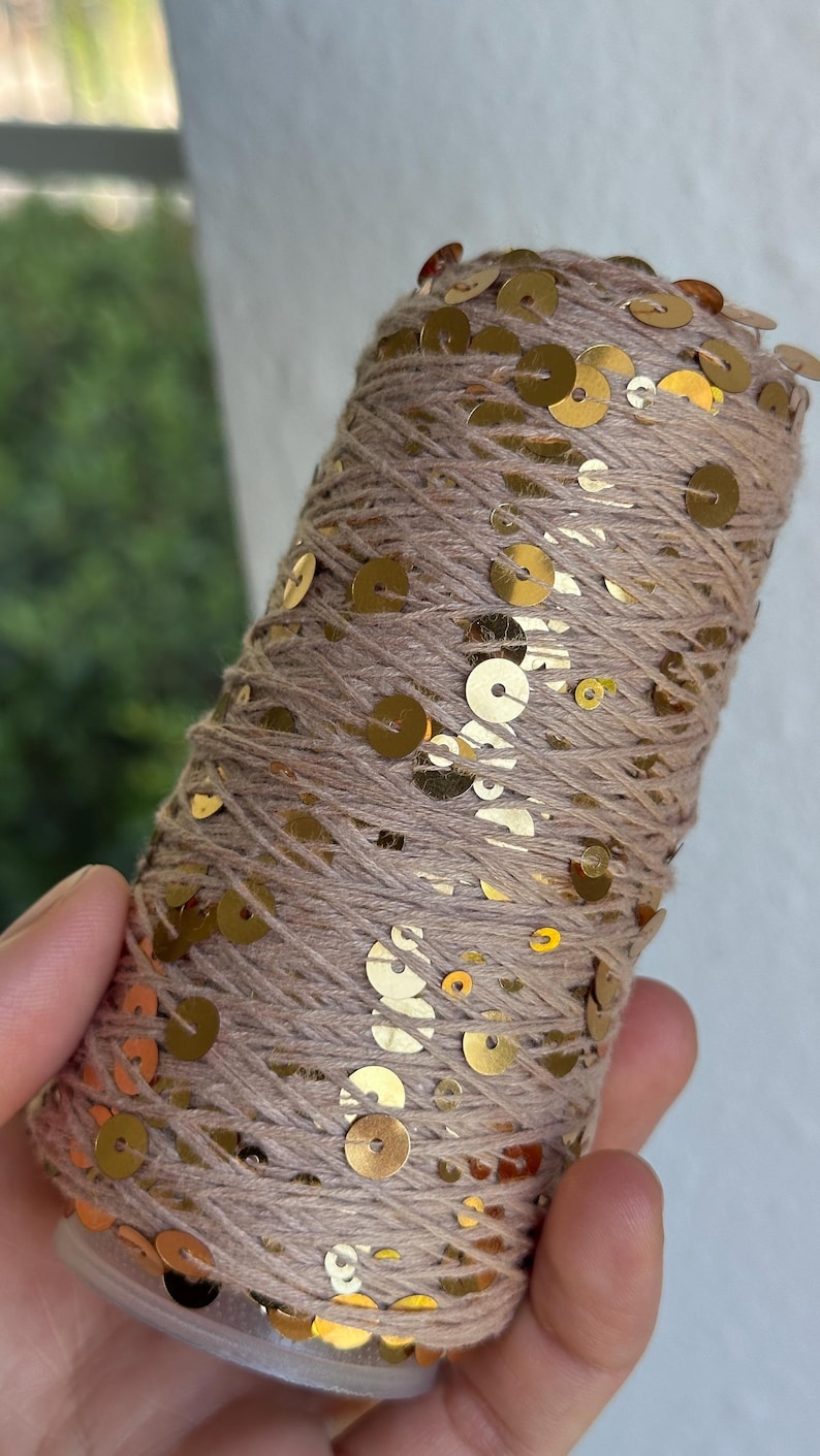May include: A cylindrical spool of light brown yarn wrapped with small, reflective gold sequins. The metallic gold discs are distributed across the surface of the wound thread, creating a shimmering texture throughout the entire length of the spool.