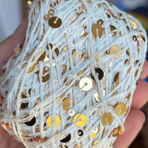 May include: A ball of white yarn interwoven with gold sequins. The yarn is loosely woven, creating a textured surface. The gold sequins are scattered throughout, adding a touch of sparkle and glamour to the craft supply.