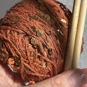 May include: A ball of rust-colored yarn interwoven with small, reflective gold sequins, paired with two light-toned wooden knitting needles. The yarn features a textured, fibrous appearance with metallic accents distributed throughout the spherical bundle.