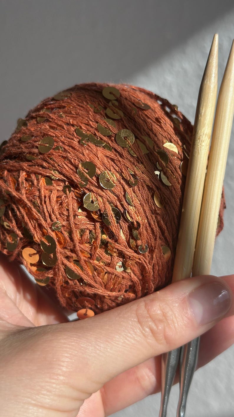May include: A ball of rust-colored yarn interwoven with small, reflective gold sequins, paired with two light-toned wooden knitting needles. The yarn features a textured, fibrous appearance with metallic accents distributed throughout the spherical bundle.