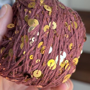 May include: A round ball of dark mauve yarn intertwined with numerous small, flat, circular gold-colored sequins. The yarn has a textured, matte appearance, while the metallic sequins are distributed throughout the surface of the ball.