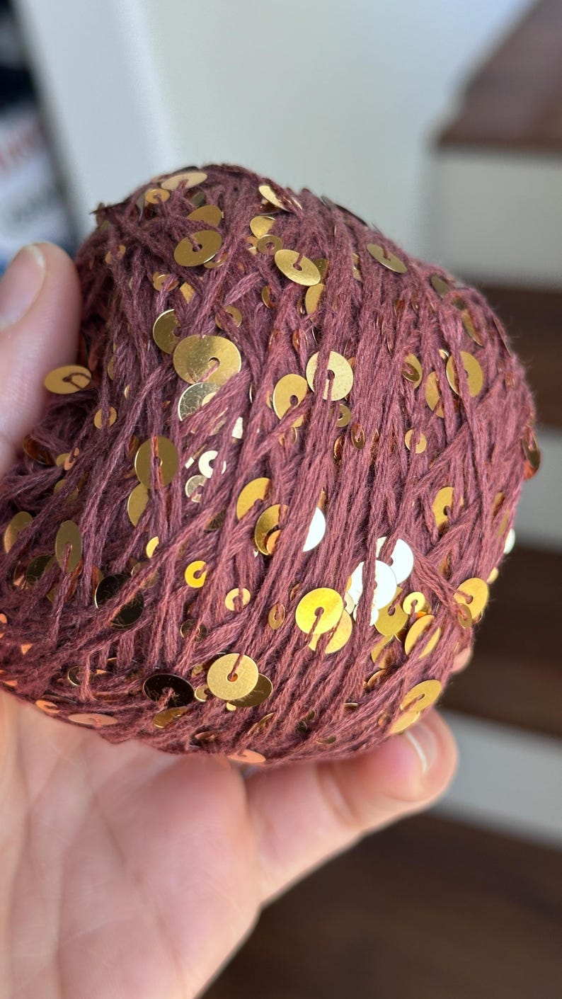 May include: A round ball of dark mauve yarn intertwined with numerous small, flat, circular gold-colored sequins. The yarn has a textured, matte appearance, while the metallic sequins are distributed throughout the surface of the ball.