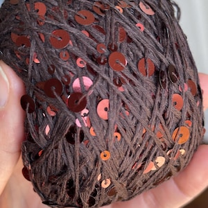 May include: A ball of dark brown yarn featuring integrated shiny copper-colored sequins. The thread is wound into a round shape, showcasing the contrast between the matte dark brown fibers and the reflective metallic circular sequins throughout.