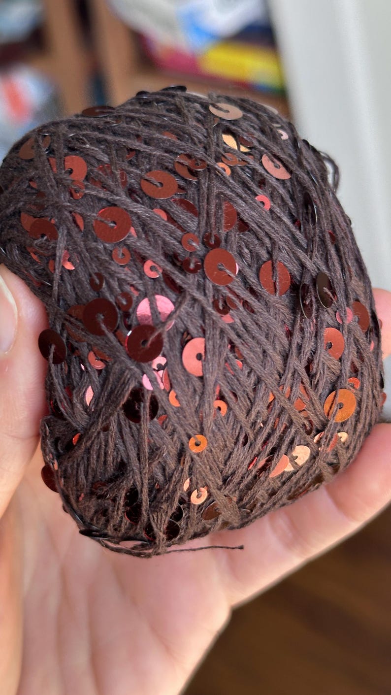 May include: A ball of dark brown yarn featuring integrated shiny copper-colored sequins. The thread is wound into a round shape, showcasing the contrast between the matte dark brown fibers and the reflective metallic circular sequins throughout.
