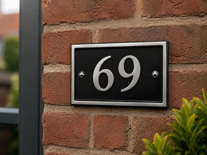 Puede incluir: Placa rectangular negra con el n&uacute;mero 69 en plata. La placa tiene un borde plateado y est&aacute; fijada a una pared de ladrillo rojo. Se ven dos remaches plateados.