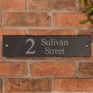 May include: A rectangular black slate house number sign with the number "2" and the words "Sulivan Street" in a light gray font. The sign is mounted on a red brick wall with two silver rivets.