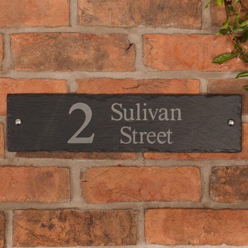 May include: A rectangular black slate house number sign with the number "2" and the words "Sulivan Street" in a light gray font. The sign is mounted on a red brick wall with two silver rivets.