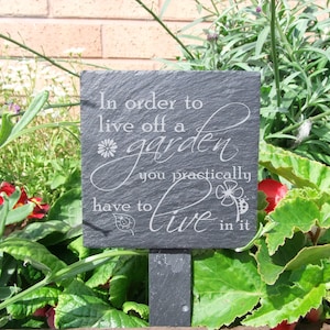 Może przedstawiać: Ciemnoszary, łupkowy znak ogrodowy z cytatem "In order to live off a garden you practically have to live in it" w białym skrypcie. Znak otoczony jest zielonymi liśćmi i kolorowymi kwiatami.