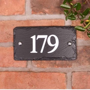 以下が含まれることがあります： 黒いスレートの長方形の家番号プレートで、白い数字の「179」が表示されています。 プレートは赤いレンガの壁に取り付けられ、2つの銀色のリベットで固定されています。 右上隅には緑の葉が見えます。