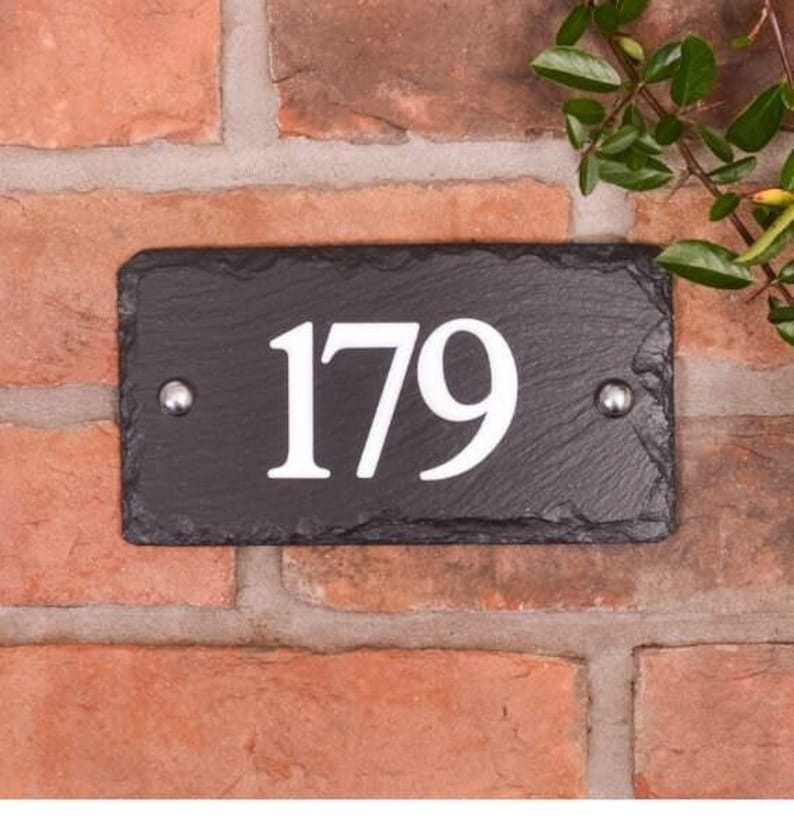 以下が含まれることがあります： 黒いスレートの長方形の家番号プレートで、白い数字の「179」が表示されています。 プレートは赤いレンガの壁に取り付けられ、2つの銀色のリベットで固定されています。 右上隅には緑の葉が見えます。