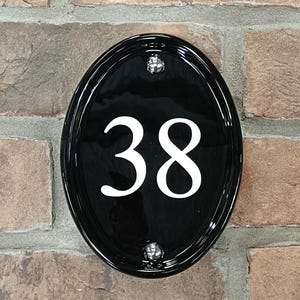 Op de afbeelding: Zwart ovale huisnummerbord met het nummer 38 in wit. Het bord is bevestigd aan een bakstenen muur en heeft twee zilverkleurige montageschroeven.