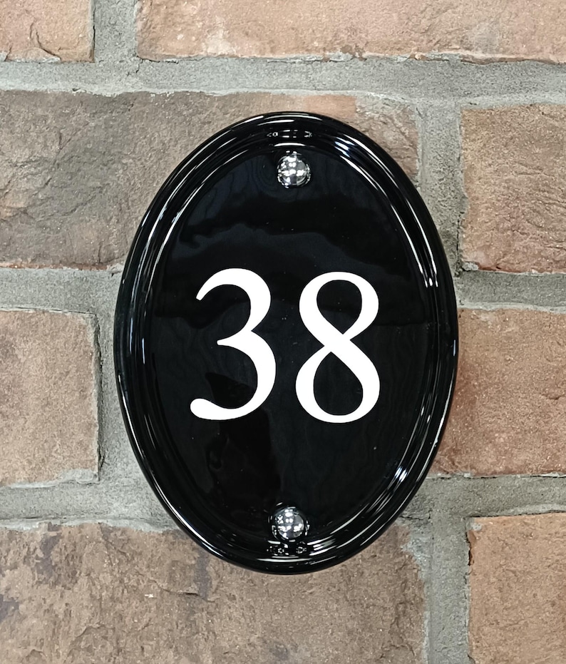 Op de afbeelding: Zwart ovale huisnummerbord met het nummer 38 in wit. Het bord is bevestigd aan een bakstenen muur en heeft twee zilverkleurige montageschroeven.
