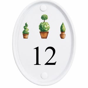 May include: White oval house number sign with the number 12 in black. The sign features three watercolor illustrations of potted topiary plants in shades of green and brown. Two white fasteners are at the top and bottom.