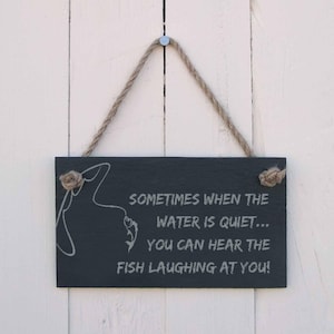 Könnte beinhalten: Rechteckiges schwarzes Schiefertafelschild mit einem Seilaufhänger. Das Schild zeigt eine weiße Illustration einer Angelschnur und eines Fisches sowie den Text: "SOMETIMES WHEN THE WATER IS QUIET... YOU CAN HEAR THE FISH LAUGHING AT YOU!"