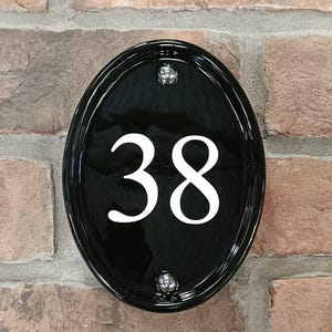 Op de afbeelding: Zwart ovale huisnummerbord met het witte nummer 38. Het bord is bevestigd aan een bakstenen muur met twee zilveren schroeven. Het glanzende oppervlak reflecteert licht.