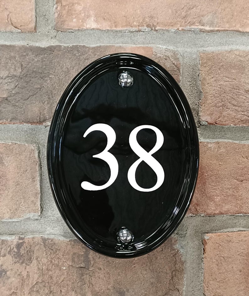 Op de afbeelding: Zwart ovale huisnummerbord met het witte nummer 38. Het bord is bevestigd aan een bakstenen muur met twee zilveren schroeven. Het glanzende oppervlak reflecteert licht.