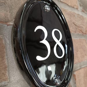 Op de afbeelding: Zwarte ovale huisnummerplaat met het witte nummer 38. De plaat is bevestigd aan een bakstenen muur en heeft een glanzende afwerking met twee zilverkleurige klinknagels.