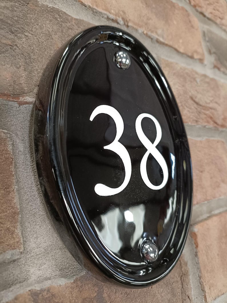 Op de afbeelding: Zwarte ovale huisnummerplaat met het witte nummer 38. De plaat is bevestigd aan een bakstenen muur en heeft een glanzende afwerking met twee zilverkleurige klinknagels.