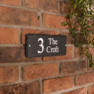May include: A rectangular black slate house number sign with the number "3" and the words "The Croft" in white lettering. The sign is mounted on a red brick wall with two silver screws.