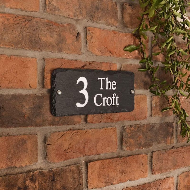 May include: A rectangular black slate house number sign with the number "3" and the words "The Croft" in white lettering. The sign is mounted on a red brick wall with two silver screws.