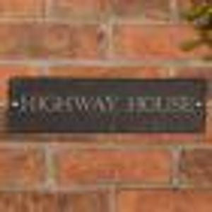 May include: A rectangular, dark gray slate sign with the text "HIGHWAY HOUSE" in white lettering. The sign is mounted on a red brick wall, with the text centered horizontally.