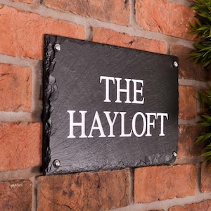 May include: A rectangular black slate sign with the words "THE HAYLOFT" in white lettering. The sign is mounted on a red brick wall with silver-colored screws in each corner.