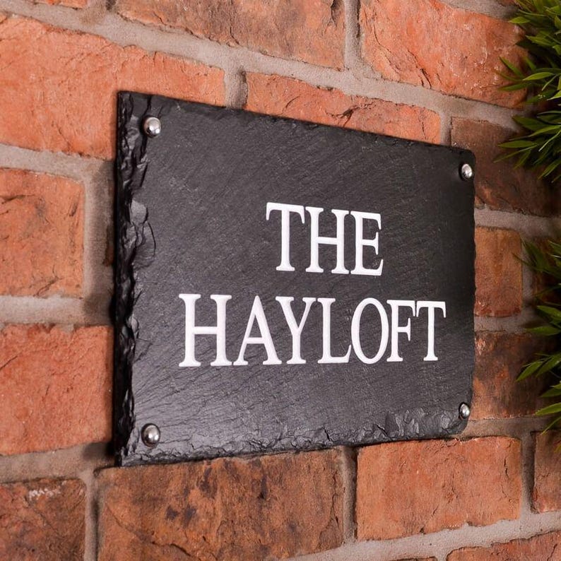 May include: A rectangular black slate sign with the words "THE HAYLOFT" in white lettering. The sign is mounted on a red brick wall with silver-colored screws in each corner.