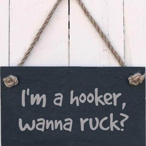 Könnte beinhalten: Rechteckiges schwarzes Schiefertafelschild mit dem Text "I'm a hooker, wanna ruck?" in weißer Schrift. Das Schild ist an einem Naturseil aufgehängt und auf einem weißen Holzhintergrund montiert.