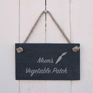 Könnte beinhalten: Rechteckiges Schild aus schwarzem Schiefer mit der Aufschrift "Mum's Vegetable Patch" und einer Karottenillustration. Es hängt an einem Naturseil und ist auf einem weißen Holzgrund befestigt.