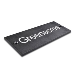 May include: A rectangular, dark grey sign with the word "Greenacres" in white, centred. The sign has a textured surface and two silver-coloured rivets. Likely for a home or business.