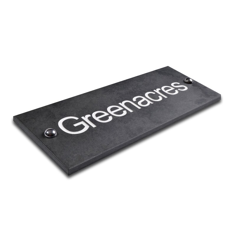 May include: A rectangular, dark grey sign with the word "Greenacres" in white, centred. The sign has a textured surface and two silver-coloured rivets. Likely for a home or business.