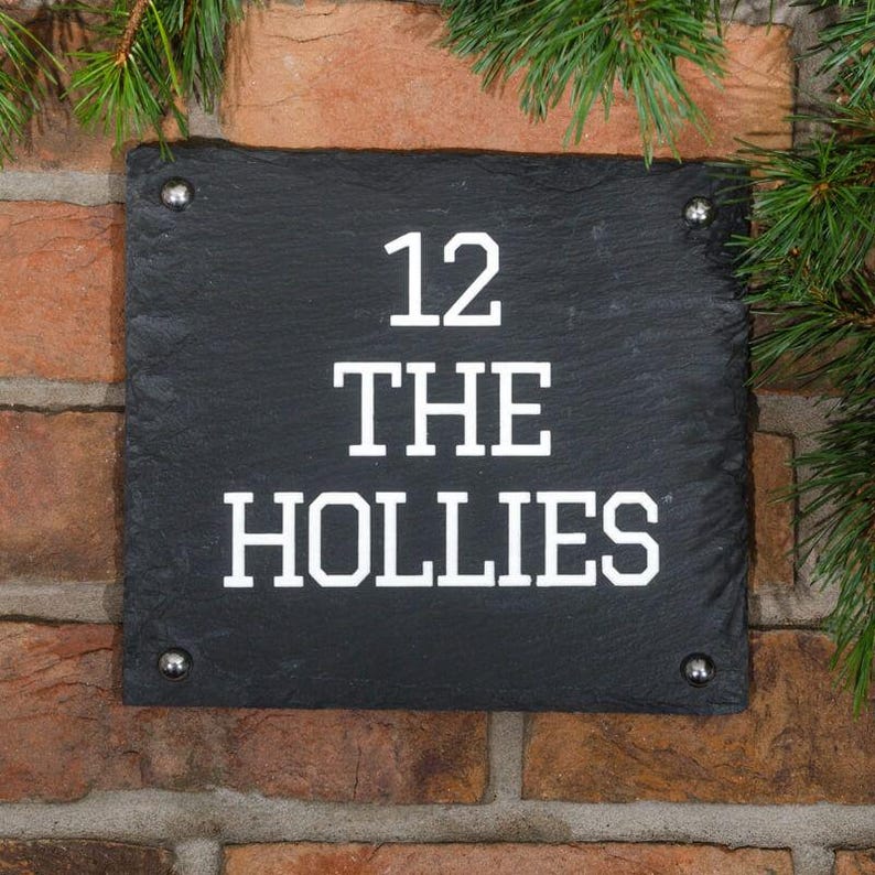 May include: A black slate house number sign with the number 12 and the name "THE HOLLIES" in white, mounted on a brick wall. The sign is rectangular with rounded corners and is secured with four silver screws.