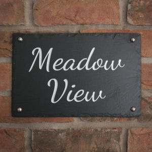 May include: A rectangular black slate sign with the words "Meadow View" in white, cursive lettering. The sign is mounted on a red brick wall with four silver-colored screws.