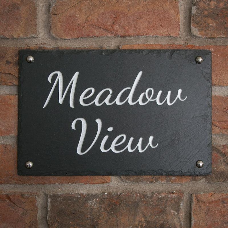 May include: A rectangular black slate sign with the words "Meadow View" in white, cursive lettering. The sign is mounted on a red brick wall with four silver-colored screws.