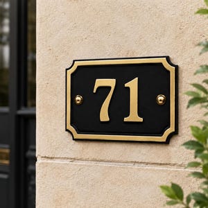 Może przedstawiać: Czarna, prostokątna tablica z numerem 71 w złotym kolorze i złotą obwódką. Tablica jest zamontowana na beżowej ścianie z tynku. W tle ciemne drzwi i liście.