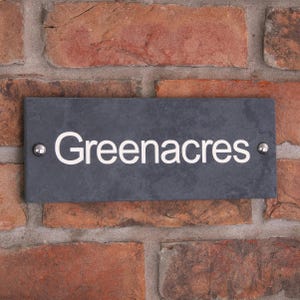 May include: A rectangular, dark grey slate sign displaying the name "Greenacres" in white lettering. The sign is fixed to a red brick wall with two silver rivets.