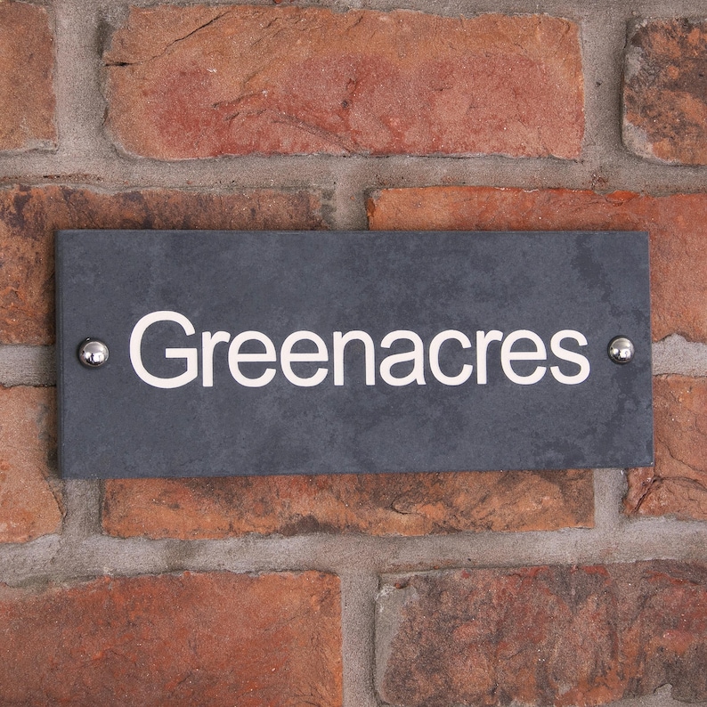 May include: A rectangular, dark grey slate sign displaying the name "Greenacres" in white lettering. The sign is fixed to a red brick wall with two silver rivets.