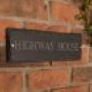 May include: A rectangular black sign with the words "HIGHWAY HOUSE" in white lettering is mounted on a brick wall. The sign is slightly weathered, suggesting outdoor use. The brick wall has a reddish-brown color.